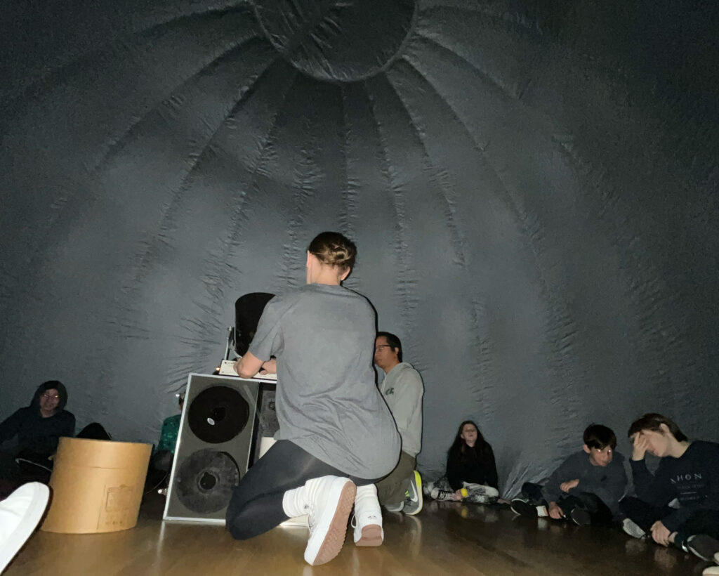 STEM Specialist Molli Grady helps prepare the StarLab projector. Students sit on the outer edge of the StarLab's interior.